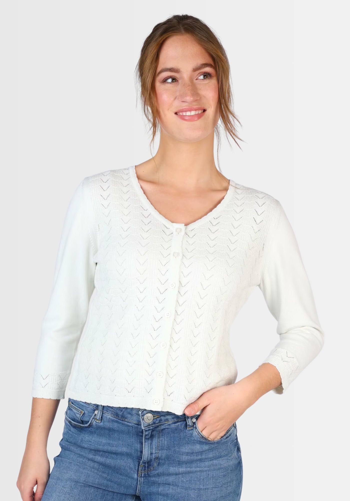 Model trägt die kurze Strickjacke „Liselotte“ in strahlendem Weiß, gefertigt aus 100% Bio-Baumwolle. Das feine Ajourmuster, der klassische Rundhalsausschnitt und die elegante Knopfleiste machen diesen Cardigan zum perfekten Begleiter zu Kleidern und Röcken.